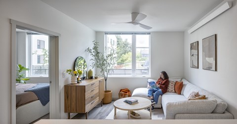 A woman is sitting on a couch in a living room with a bed, a table, and a plant.