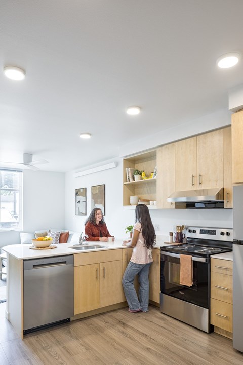 A woman is standing in a modern kitchen with wooden cabinets and stainless steel appliances.
