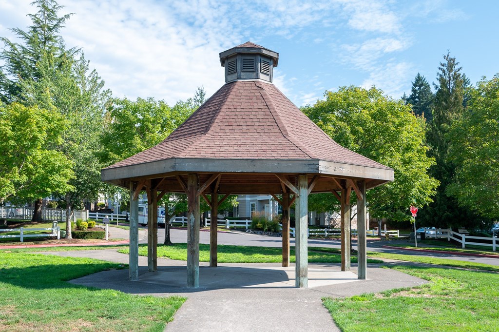 A gazebo with a brown roof is surrounded by a grassy area and trees.