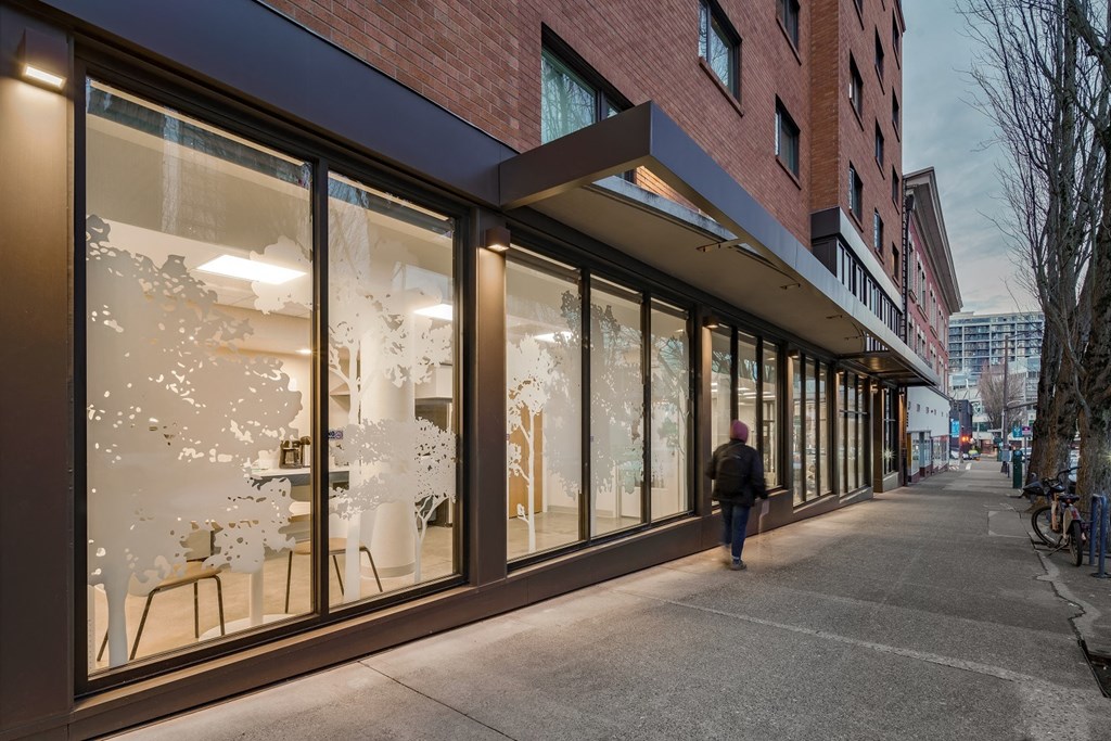 a woman walks down a sidewalk in front of a building with a large window with a tree