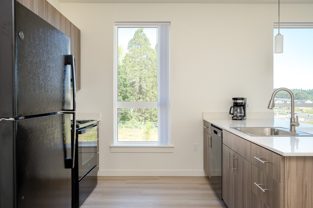 A black fridge stands in a kitchen with a window showing trees outside.
