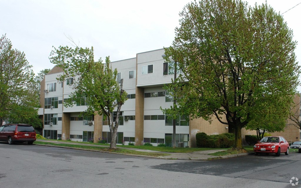 A red car is parked on the street in front of a white apartment building.