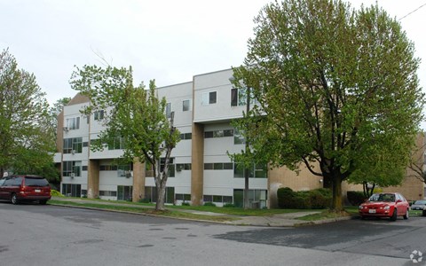 A red car is parked on the street in front of a white apartment building.