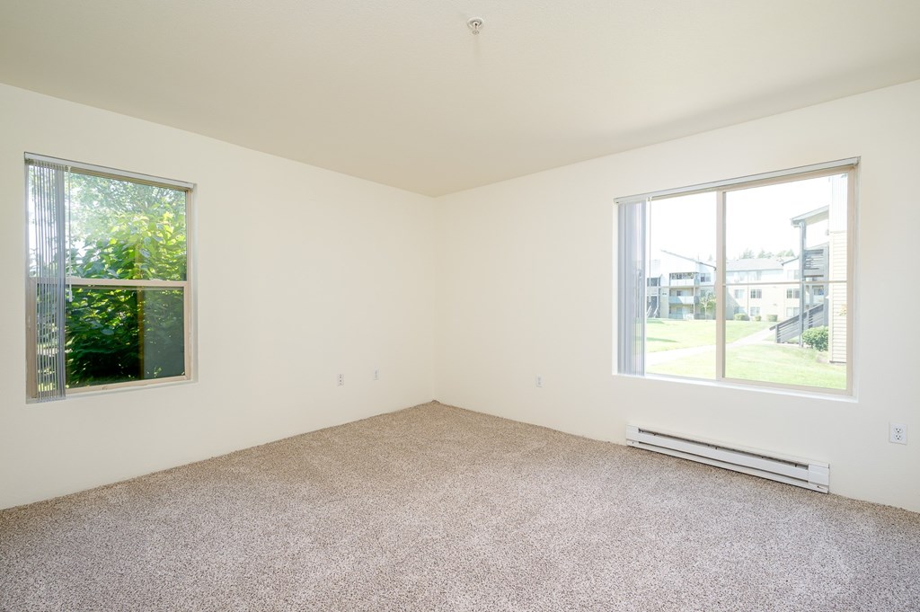 A room with a carpet floor, a window with a view of a building and trees outside, and a wall-mounted air conditioner.
