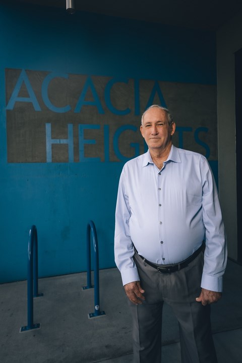 A man stands in front of a blue wall with the words "ACACIA HEIGHTS" on it.