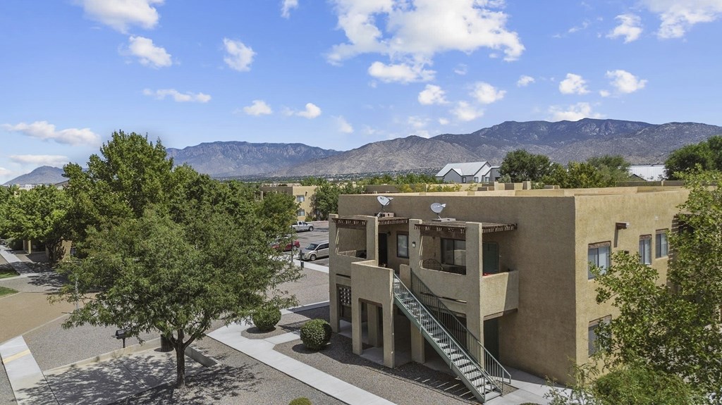A modern building with a staircase leading to the entrance is surrounded by trees and has mountains in the background.