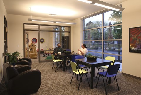 A conference room with a long table and chairs, a person standing at the head of the table, and a view of trees outside the window.