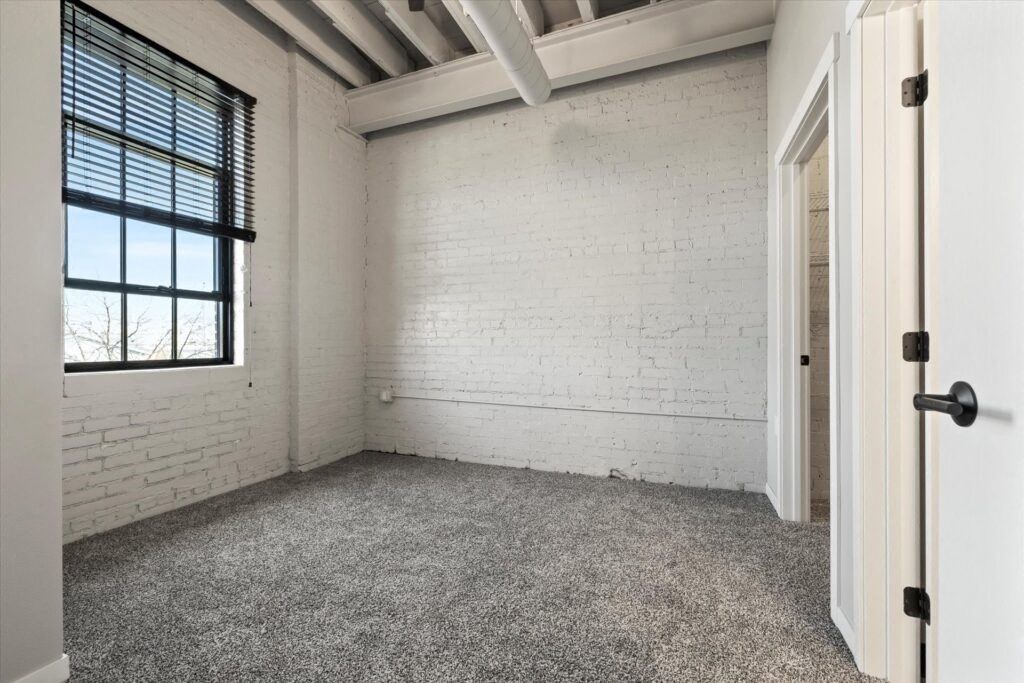 a bedroom with carpeting, a large window, and a whitewashed brick wall