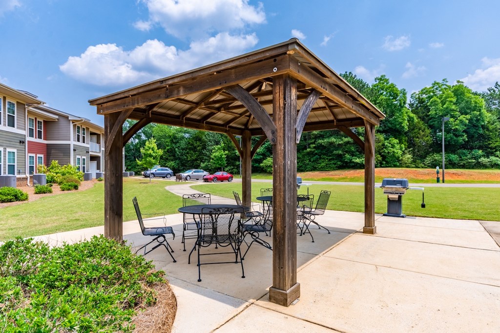 A wooden pavilion with a table and chairs is surrounded by a grassy area.