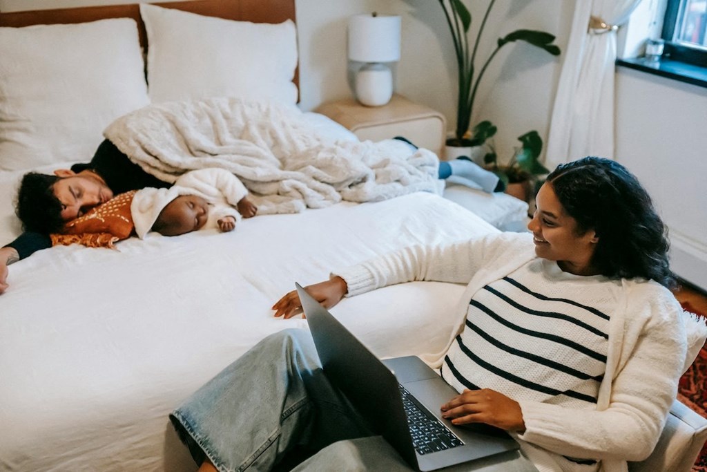 a woman sitting on a lounge with a laptop on her lap and a man and child laying on a bed
