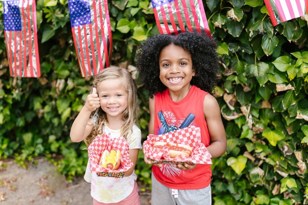 two young girls holding hot dogs in front of American flags and shrubbery