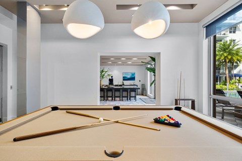 A pool table with a cue stick and balls is in the foreground of a room with a view of a dining area and outdoor patio.