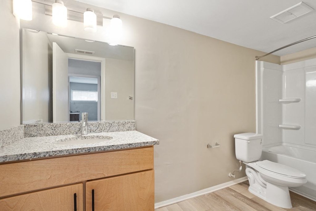 A bathroom with a granite topped vanity with mirror and light and toilet and tub/shower