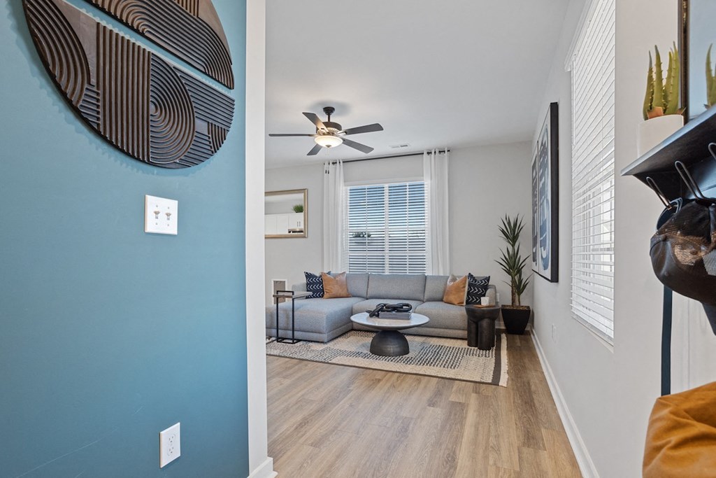 looking from a hallway into a large living room with large windows, an illuminated fan, and model furnishings
