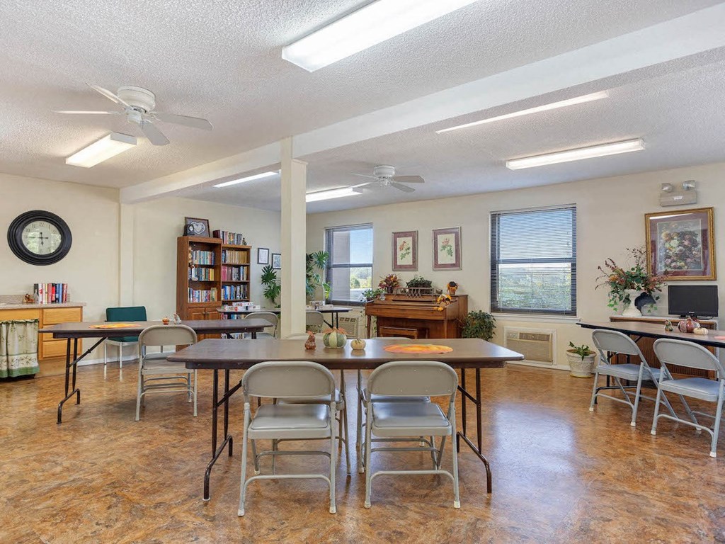 The Village Square community room with tables and chairs and a piano and bookshelves