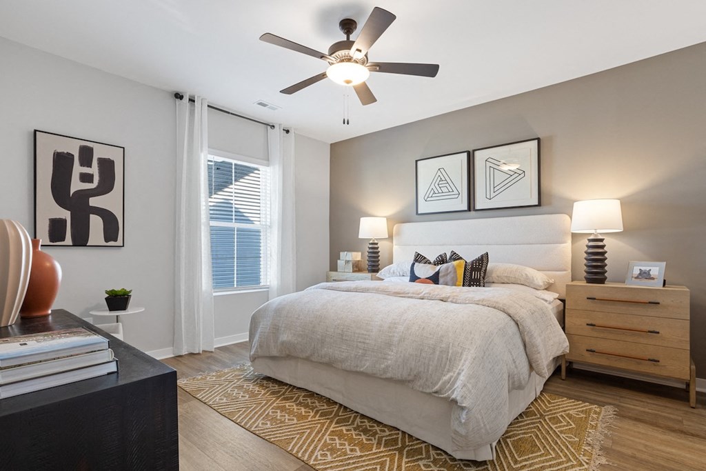 A primary bedroom with an illuminated ceiling fan, large window, model furnishings, and wood-style flooring