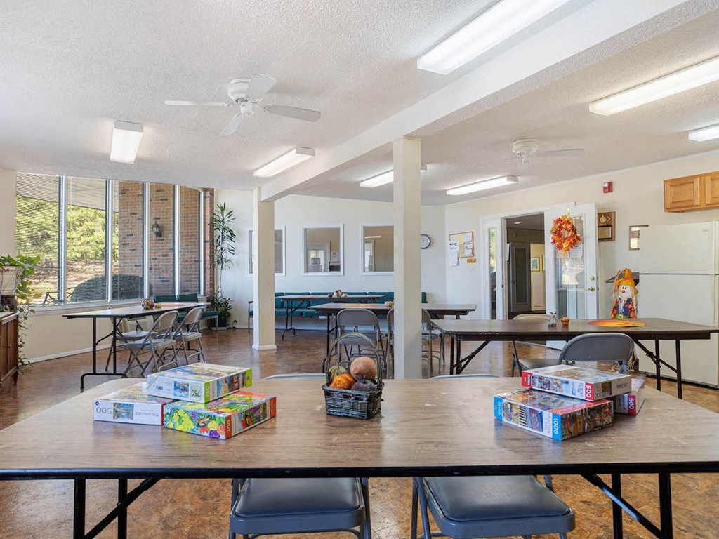 a large community room with tables, chairs, puzzles, and large windows at Village Square