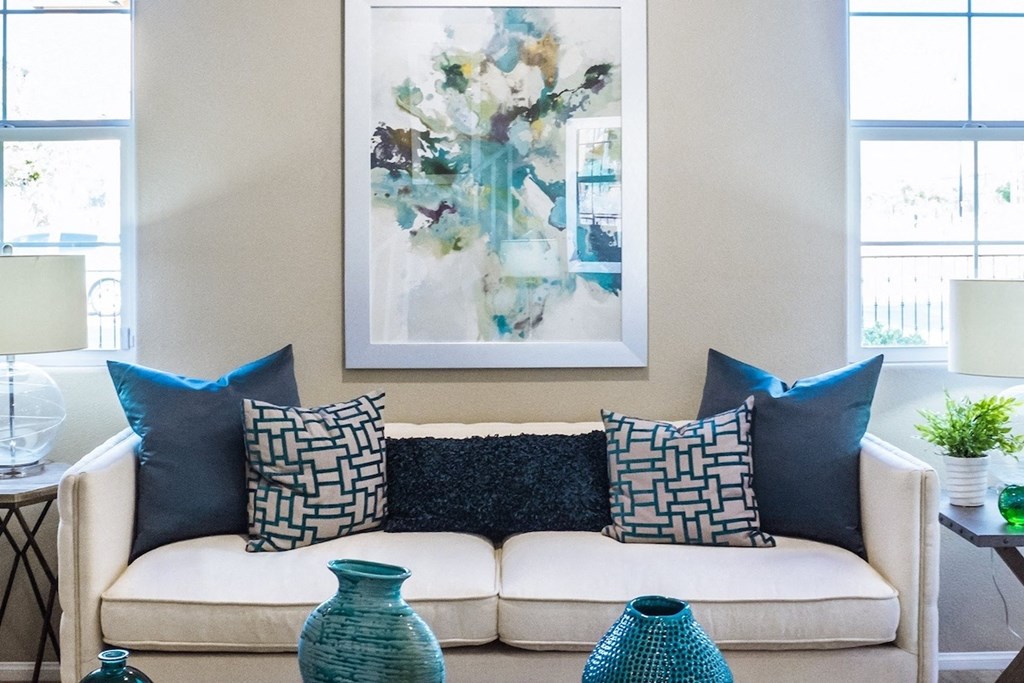 a living room with a couch and pillows and a painting on the wall flanked by windows