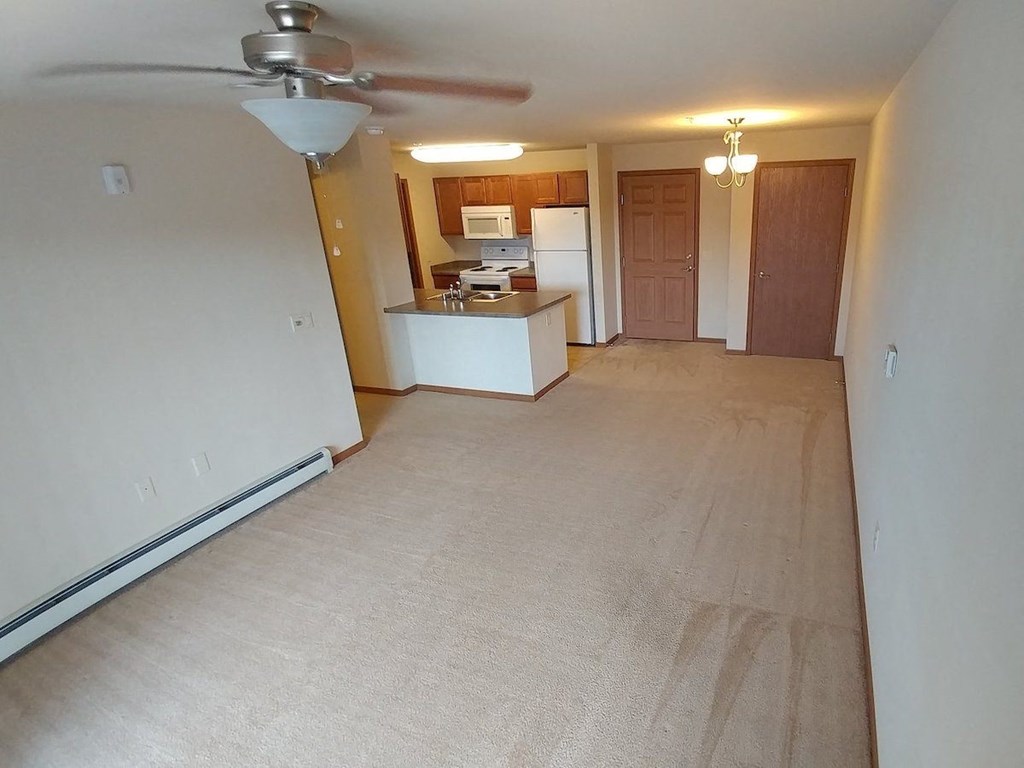 an empty living room with a ceiling fan and a kitchen in the background
