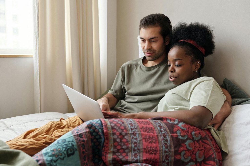 a man and a woman laying in bed looking at a laptop with natural light streaming in the window
