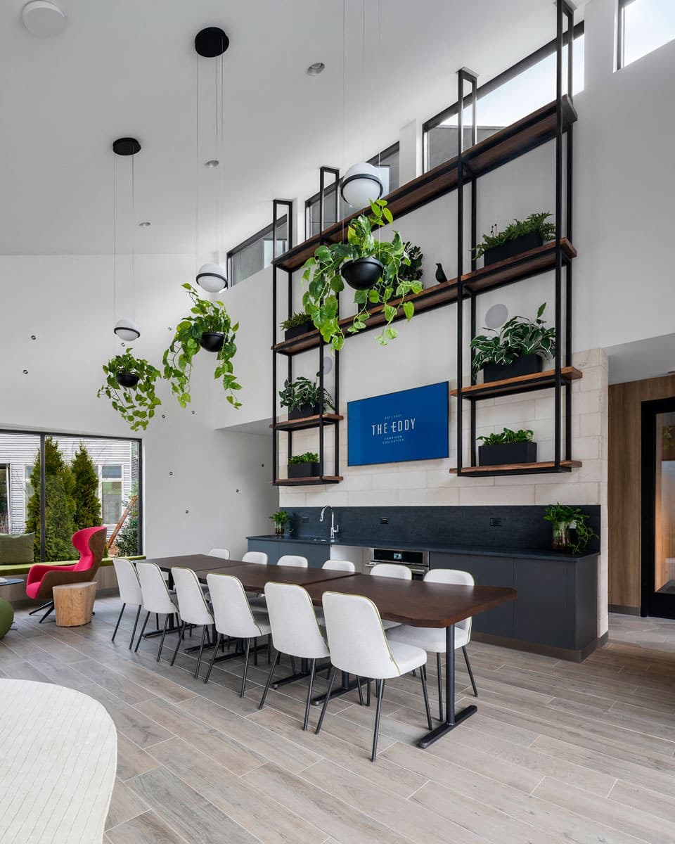 a long table with white chairs in front of a white wall with plants on it and a