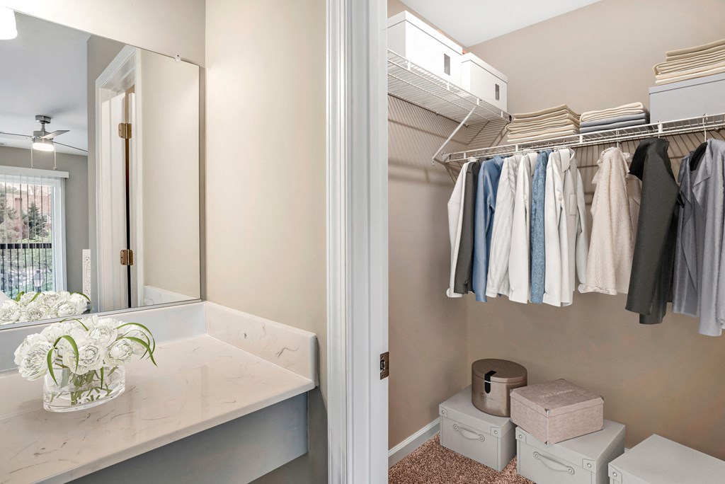A virtually staged walk-in closet with gray walls and white trim. The closet is staged with a variety of clothing, white boxes, and linens. A marble countertop vanity area with mounted mirror is located just outside of the closet.