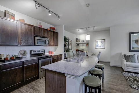 Kitchen island with granite countertop, double basin sink, hanging light fixture, barstools, dark wood cabinetry and stainless appliances