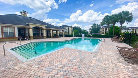 Community pool with sundeck, lounge chairs surrounded by black metal fence with palm trees, and trees, and buildings in the background