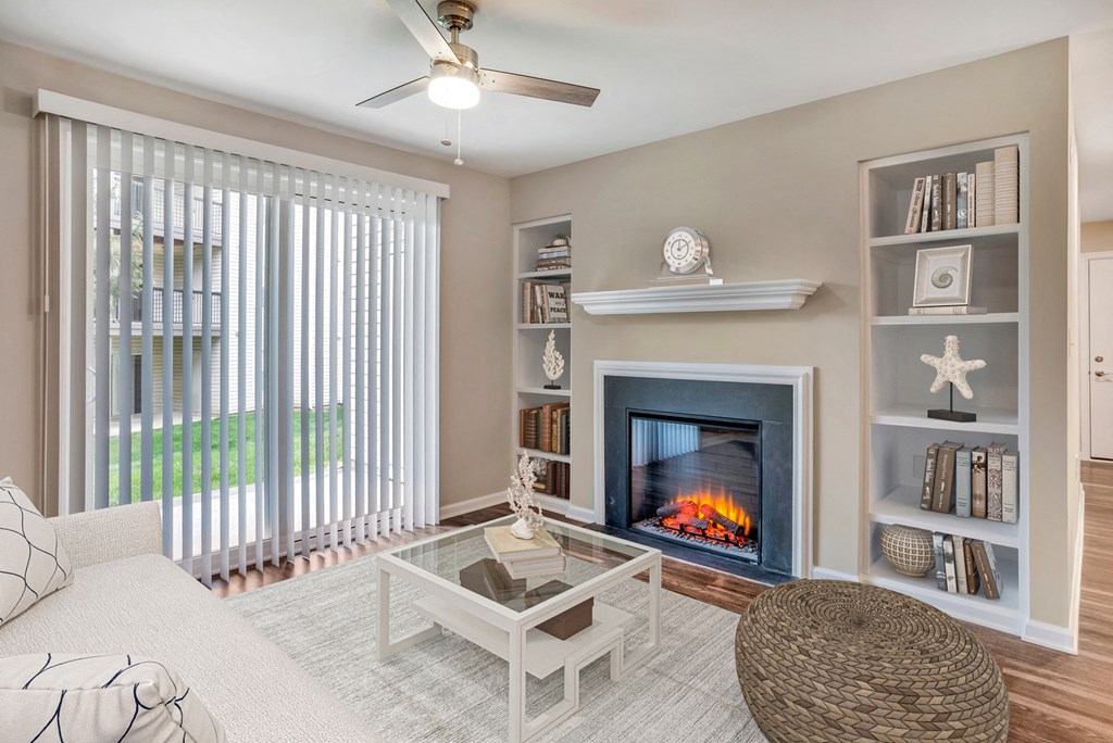 A virtually staged living room with hardwood style flooring, gray walls with white trim, a sliding glass door with vertical blinds, an electric fireplace and built-in shelves on either side of the fireplace.