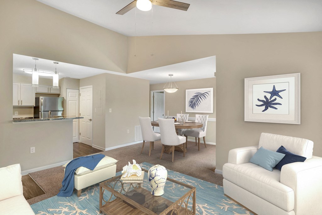A virtually staged living and dining room with gray walls, gray carpet, vaulted ceilings and a contemporary three blade ceiling fan in the living room. The living room is beach theme with a white chair, white ottoman with blue throw blanket, a light blue rug patterned with white coral, and starfish artwork hanging on the wall.
