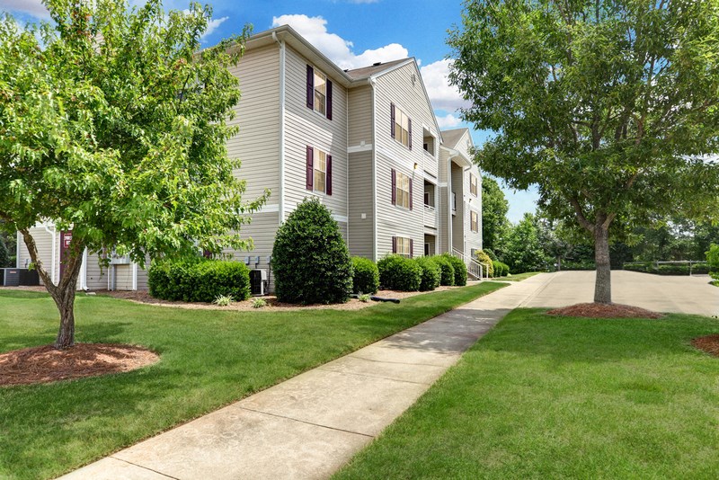 An apartment building with gray siding and burgundy shutters, is three stories tall with a balcony/patio on each level. There is a sidewalk in front of the building and the building is surrounded with grass and trees.