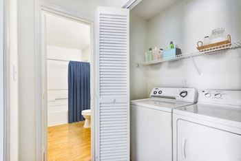 A virtually staged laundry area with accordion doors, a full size washer and dryer, white walls and built-in shelving above the washer and dryer. It is staged with two baskets and cleaning supplies. The bathroom is located in the background.