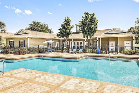 Community pool with sundeck, lounge chairs, surrounded by black metal fence with trees, lake, and exterior of clubhouse in the background