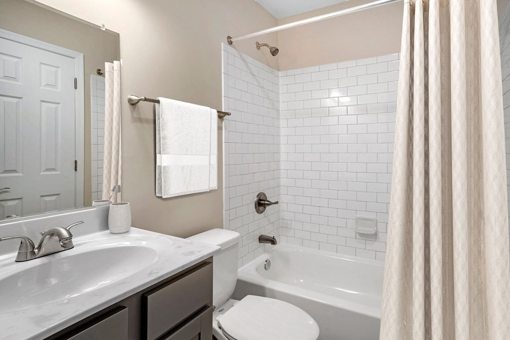 A vacant renovated bathroom with a single sink with marble countertops, gray ash cabinets below, brushed nickel plumbing fixtures, a single mounted mirror, one toilet, a towel bar, and a tub/shower combo with white subway tile surround.