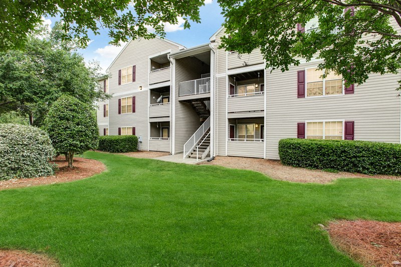 An apartment building with gray siding and burgundy shutters, is three stories tall with a balcony/patio on each level, and has stairway access in the middle of the building. The building is surrounded by grass and trees.