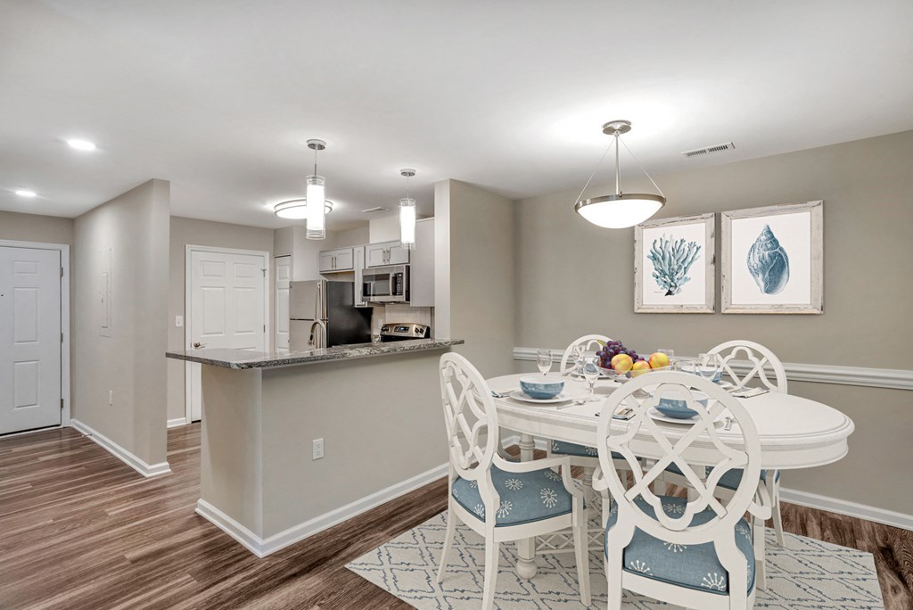 A virtually staged dining room with gray walls with white trim, hardwood style flooring and a pendant light in the center of the ceiling. A kitchen with granite countertops and stainless steel appliances is located in the background.