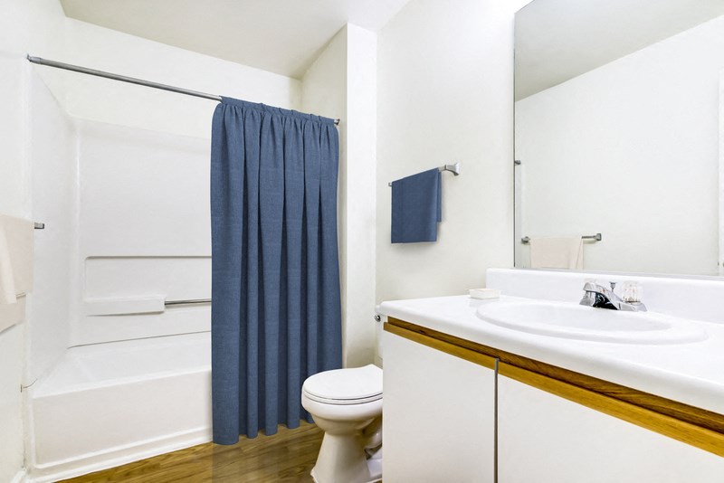 A virtually staged bathroom with hardwood style flooring, white painted walls, white countertops with single sink and white cabinets below, a single mounted mirror, one towel bar, one toilet and a shower/tub combo. It is staged with a navy blue shower curtain and matching towels