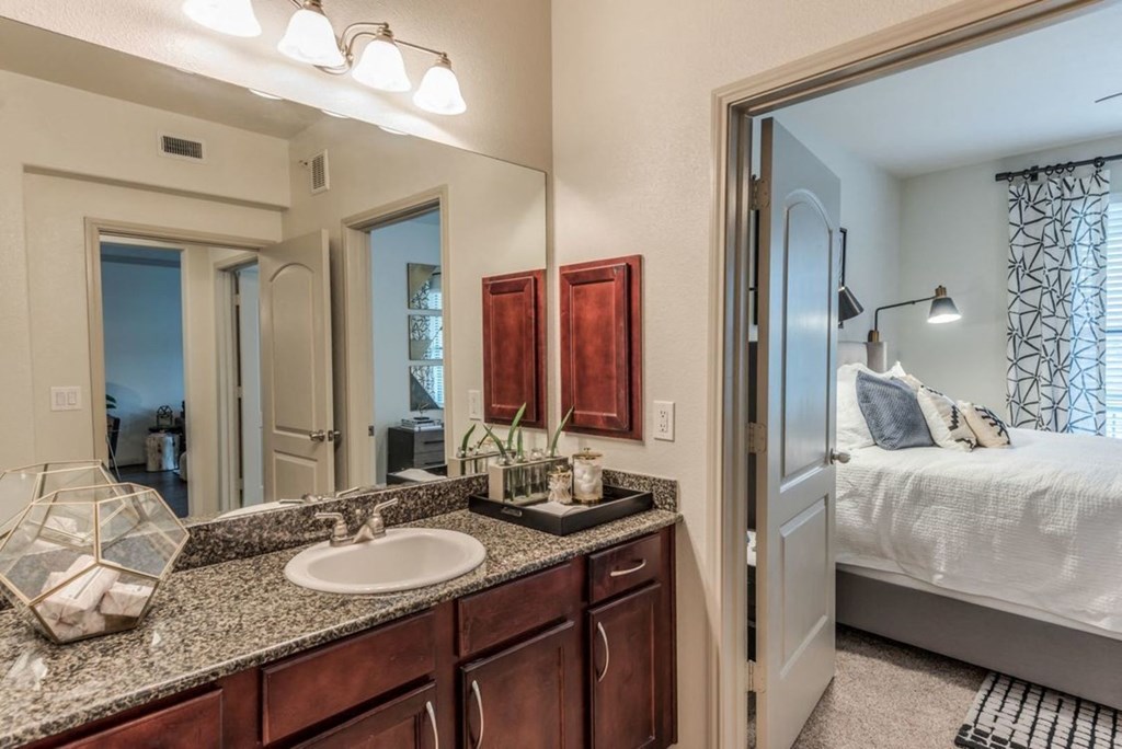 a bathroom with a sink and mirror and a bedroom in the background