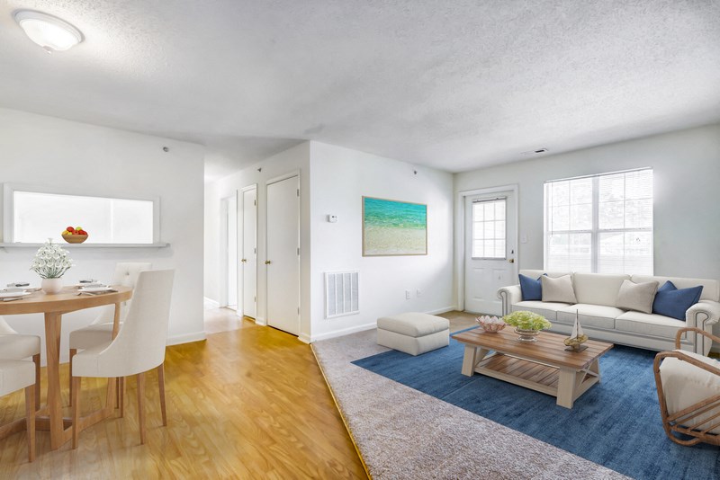 A virtually staged living room and dining room area. The living room has carpet throughout with white walls, an entry door and large picture window with blinds.