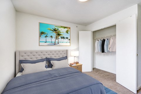 A virtually staged bedroom with white walls and gray carpet throughout, with a standard closet with double doors. It is staged with a queen gray upholstered bed with navy blue and gray bedding, a single wood night stand with lamp, and framed artwork hanging above the bed