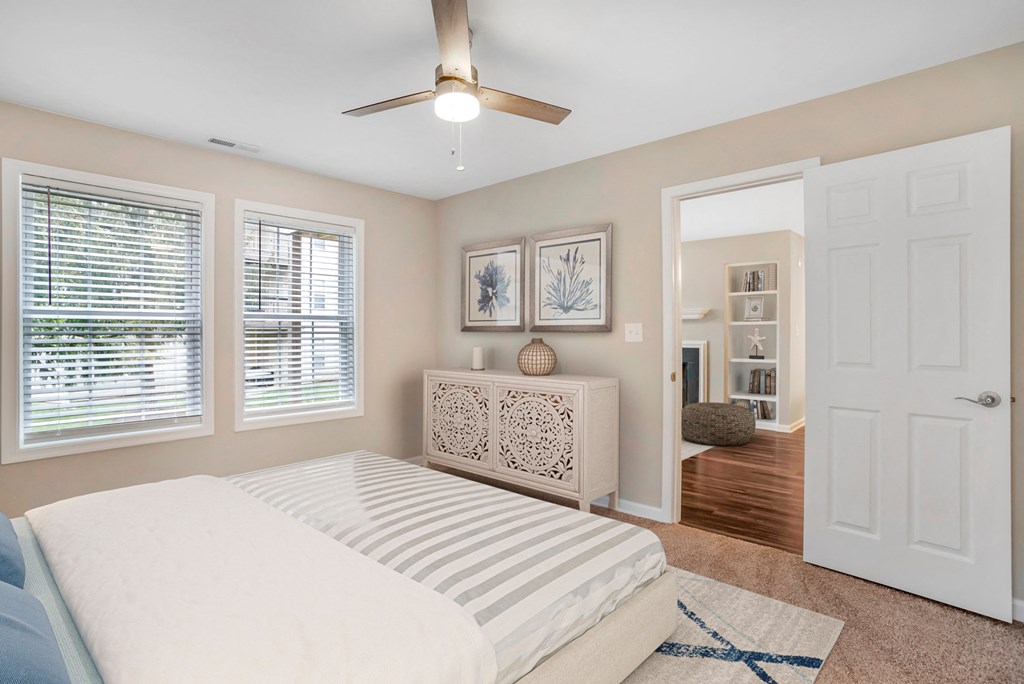 A virtually staged bedroom with the living room located in the background. The bedroom features gray walls with white trim, carpet throughout, two single windows with blinds, a three blade ceiling fan, and a door that opens to the living room.