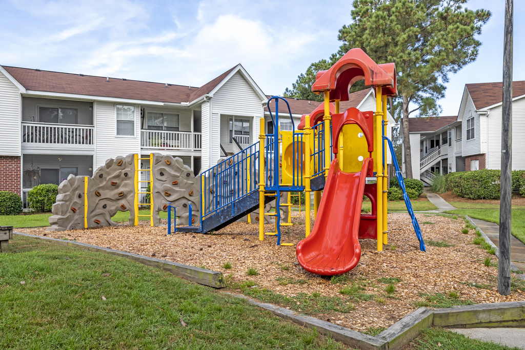 Outdoor playground with slide and climbing wall with wood chips on the ground surrounded by apartment buildings.
