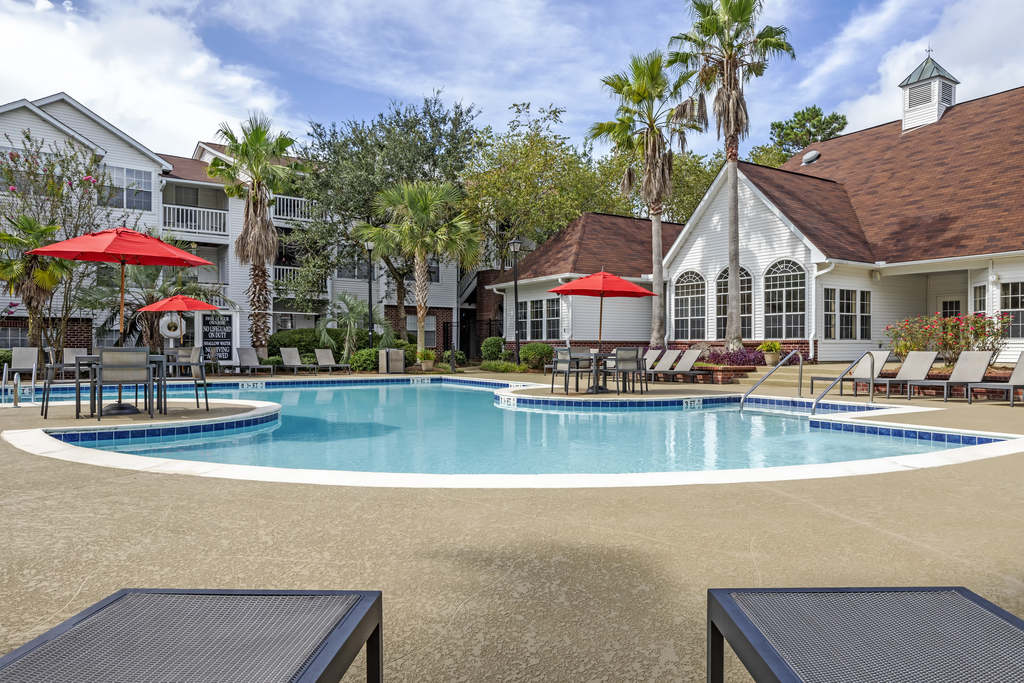 Swimming pool and sun deck with lounge chairs, tables with umbrellas, with native trees and building exteriors in the background.