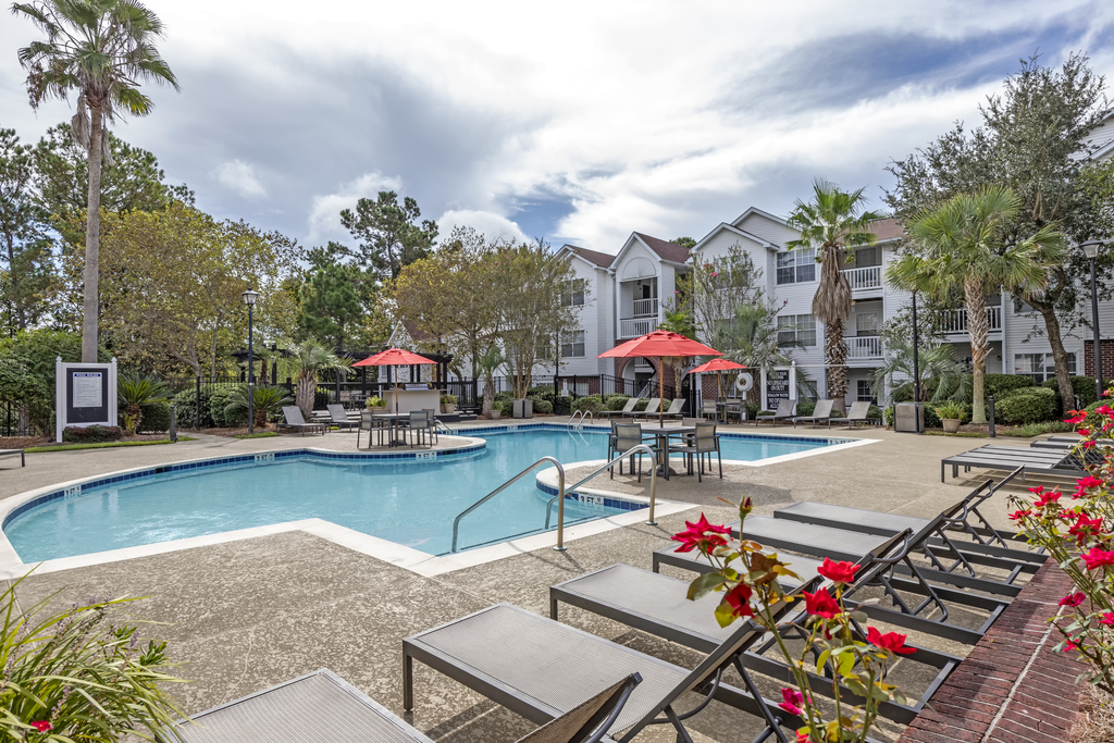 Swimming pool and sun deck with lounge chairs, tables with umbrellas, with native trees and building exteriors in the background.