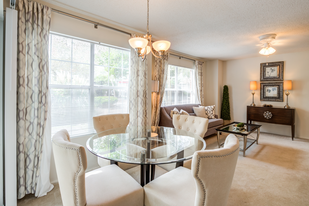 Staged dining room with white walls, carpeted floors, glass table with four chairs, decorative curtains on two large windows, and living room in the background.