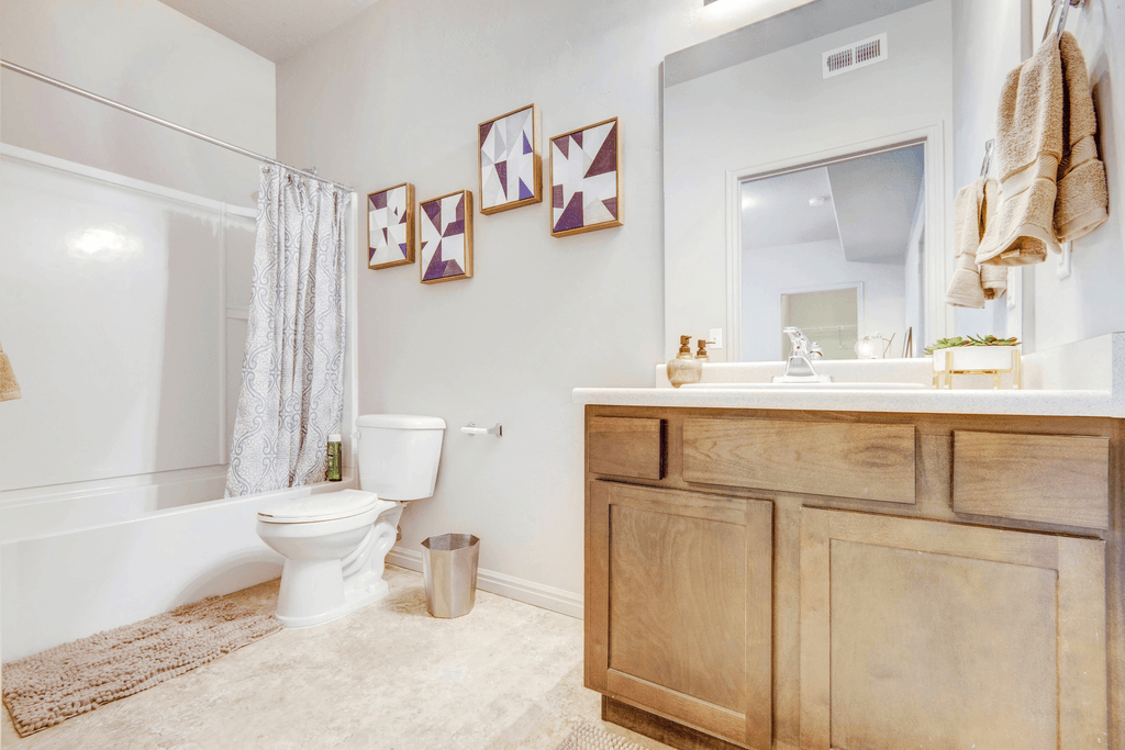 Virtually staged bathroom with tan cabinetry, large mirror, white countertop, tub with shower curtain and wall decor