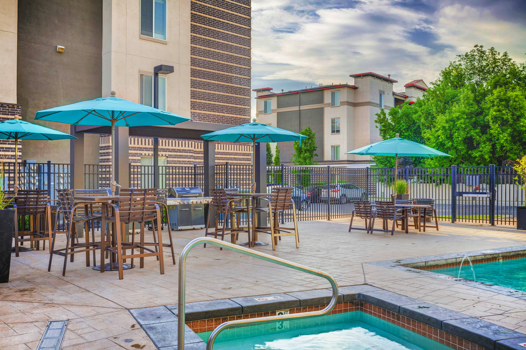 Swimming pool with heated spa, tables and chairs with blue umbrellas on the tanning deck and building exterior and native landscape surrounding.