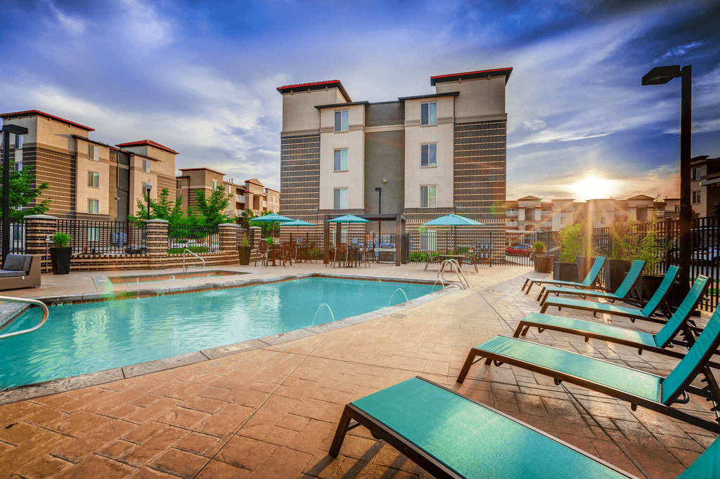 Swimming pool and heated spa with tanning deck, tables and chairs with blue umbrellas and blue lounge chairs with surrounding building exterior and native landscape.
