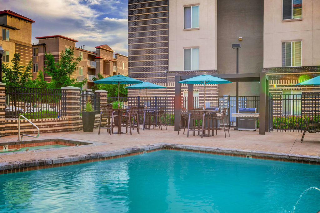 Swimming pool and heated spa with tanning deck, tables and chairs with blue umbrellas and grill area surrounded by building exterior and native landscape