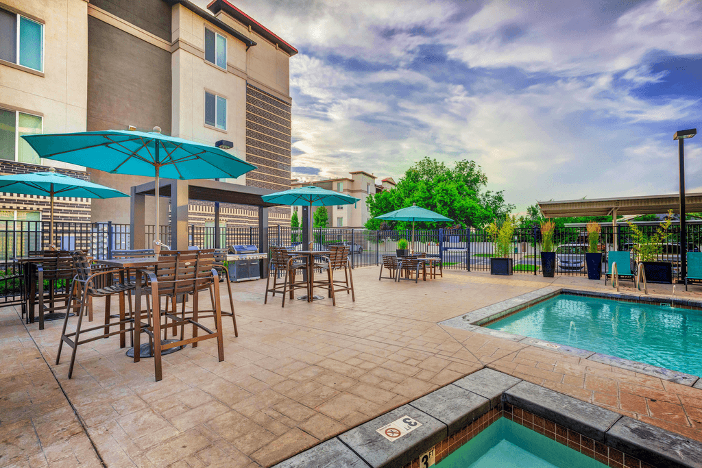 Swimming pool and heated spa with grill area in background with tanning deck, tables and chairs with blue umbrellas and surrounding native landscape and building exterior